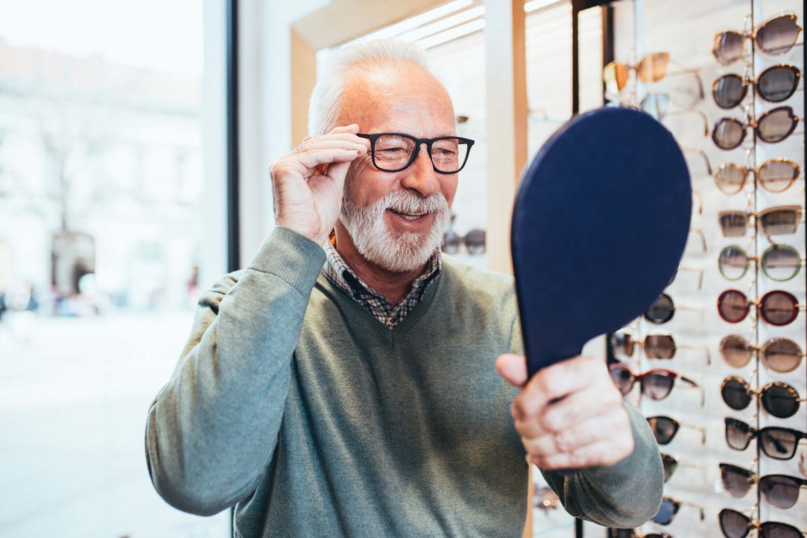 Older gentleman holding a hand-held mirror while adjusting his glasses and smiling